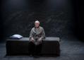An elderly man sitting on a bed. He's wearing a grey robe. The bed too is grey as is the background. The actor is Max Gillies, performing a scene from 'Endgames', a trio of short modernist plays or play extracts at Melbourne venue fortyfivedownstairs.