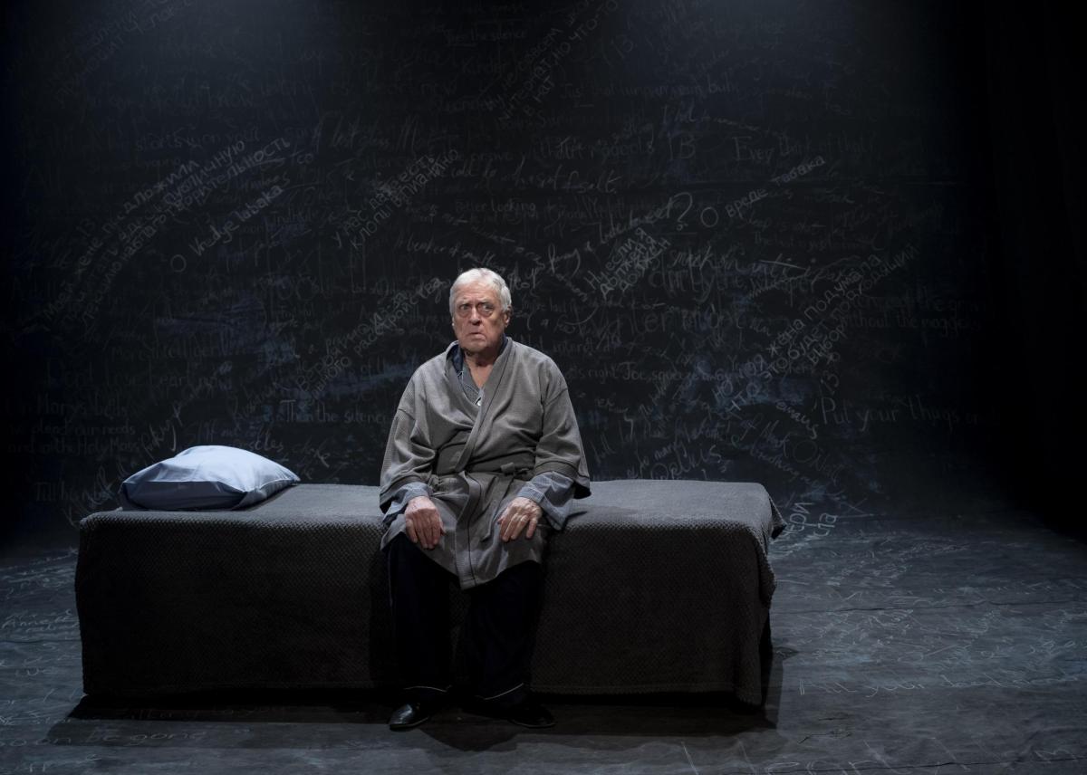 An elderly man sitting on a bed. He's wearing a grey robe. The bed too is grey as is the background. The actor is Max Gillies, performing a scene from 'Endgames', a trio of short modernist plays or play extracts at Melbourne venue fortyfivedownstairs.