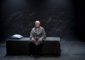 An elderly man sitting on a bed. He's wearing a grey robe. The bed too is grey as is the background. The actor is Max Gillies, performing a scene from 'Endgames', a trio of short modernist plays or play extracts at Melbourne venue fortyfivedownstairs.