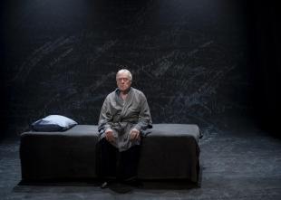 An elderly man sitting on a bed. He's wearing a grey robe. The bed too is grey as is the background. The actor is Max Gillies, performing a scene from 'Endgames', a trio of short modernist plays or play extracts at Melbourne venue fortyfivedownstairs.