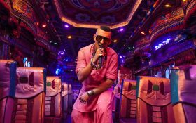 A rapper dressed in pink matching top and pants holding a microphone inside an elaborately decorated bus.