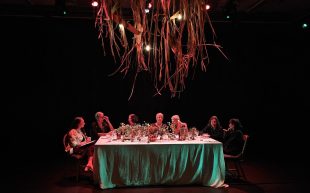 A dinner table set on a small theatre stage with six First Nations women plus one host sitting down and having conversations.