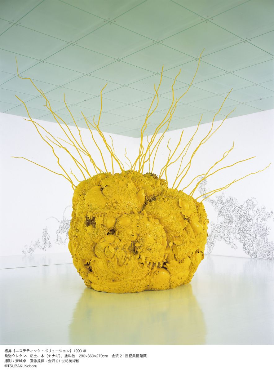 A large yellow sculpture with organic forms that appear like yellow moss, and thin branches shooting out from the top, inside a white gallery space.