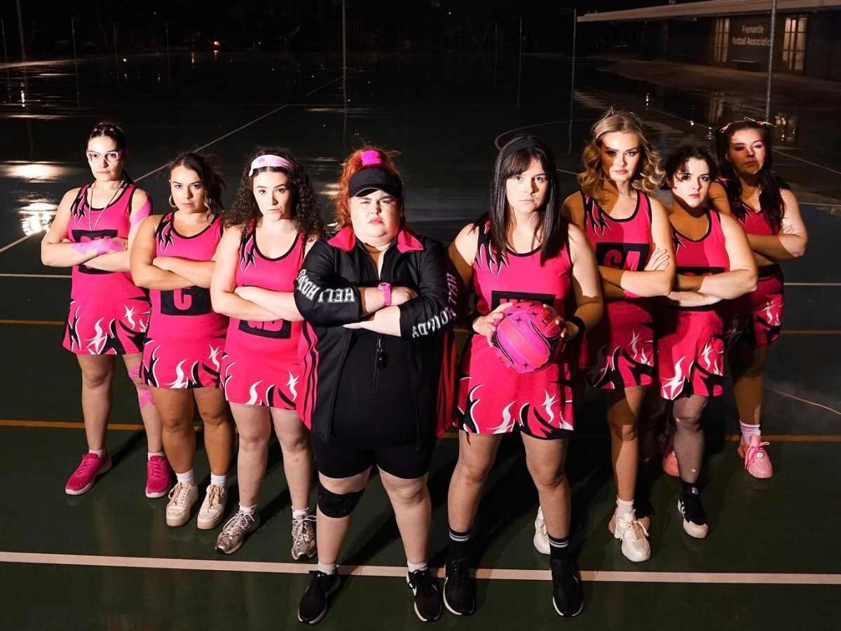 A girl’s netball team dressed in pink uniform posing for the camera looking tough on a dark sporting field. The show travels to Edinburgh.