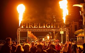 Entrance to an outdoor festival at night with fire on both sides of a large signage with the words ‘FIRELIGHT’.
