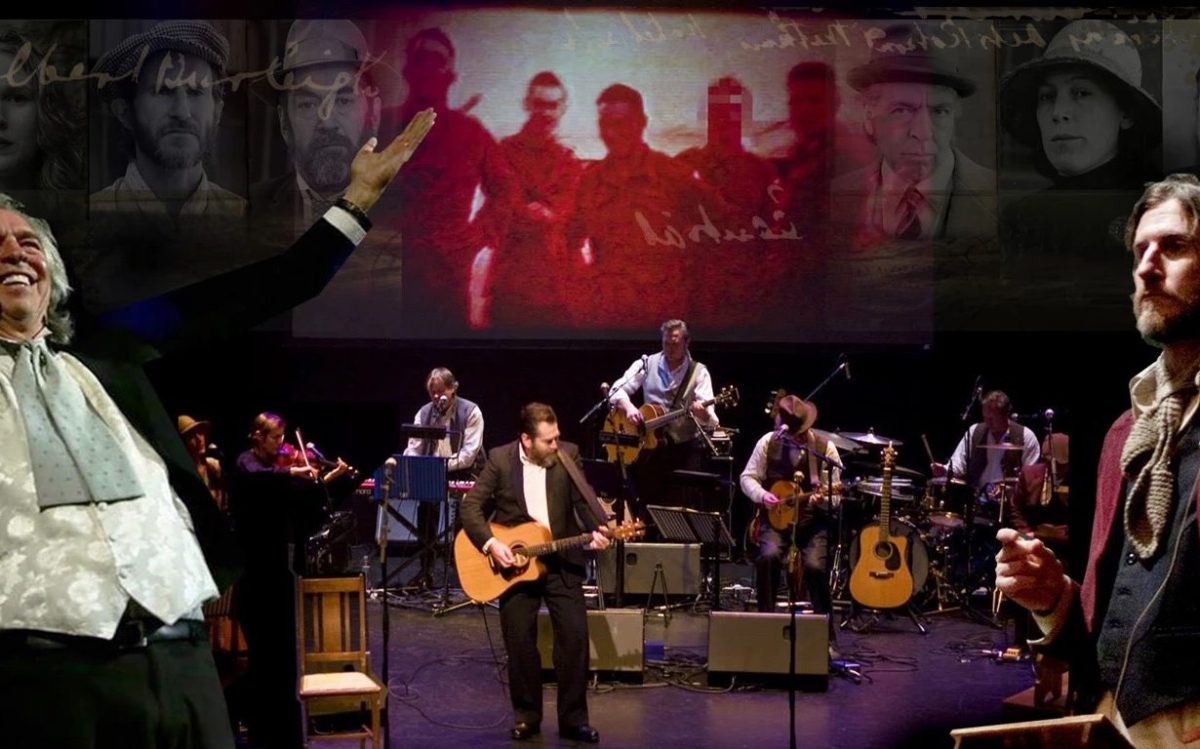 A band on stage with a video screen behind them. There are two men standing on either side of the frame.