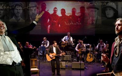 A band on stage with a video screen behind them. There are two men standing on either side of the frame.