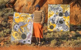 A First Nations Elder standing confidently outdoors in the red desert with two of her large-scale paintings. The paintings feature large circles, dots, and washes of paint on yellow, gray and black.