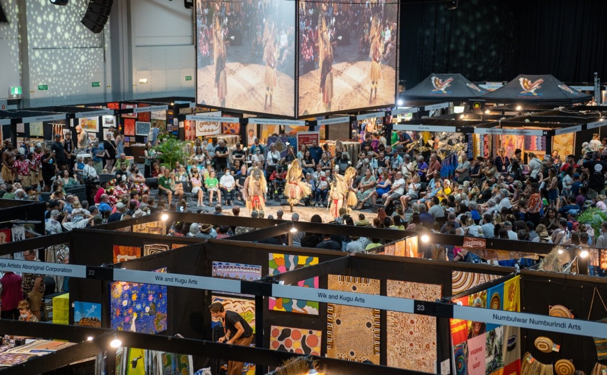 A photo taken inside a convention centre with many art stalls and a packed audience. At the centre of the image is an open space where First Nations dancers are in the middle of a performance. Photo supplied by DAAF.