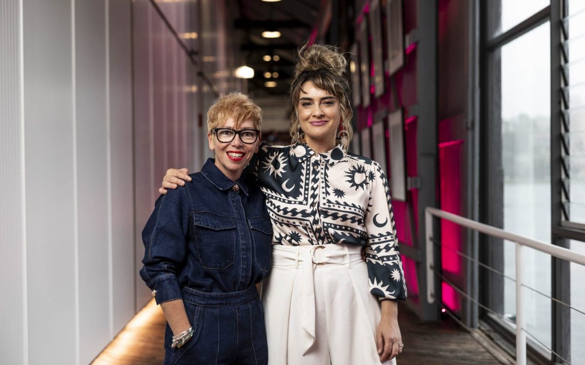 Two woman holding each other by the shoulders with warm smiles. On the left is Karolina Ristevski, a middle-aged woman wearing black-rimmed glasses and red lipstick, and on the right is Sheridan Harbridge, a younger woman with her hair in an up-do and wearing a patterned black and white top. They appear to be standing in a corridor with pink lights behind them.