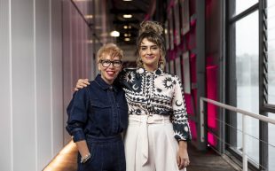 Two woman holding each other by the shoulders with warm smiles. On the left is Karolina Ristevski, a middle-aged woman wearing black-rimmed glasses and red lipstick, and on the right is Sheridan Harbridge, a younger woman with her hair in an up-do and wearing a patterned black and white top. They appear to be standing in a corridor with pink lights behind them.