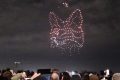 A photo of a gray-blue night sky with drones forming the outlines of a fox head. Some audiences are taking out their phones to capture a photo in the rows in front.