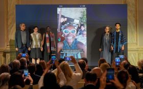 A group of five people (Venice Biennale 2026 curatorial team) standing on stage at a press conference with a projection of Koyo taking a selfie with a big smile showing between them.