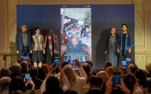 A group of five people (Venice Biennale 2026 curatorial team) standing on stage at a press conference with a projection of Koyo taking a selfie with a big smile showing between them.