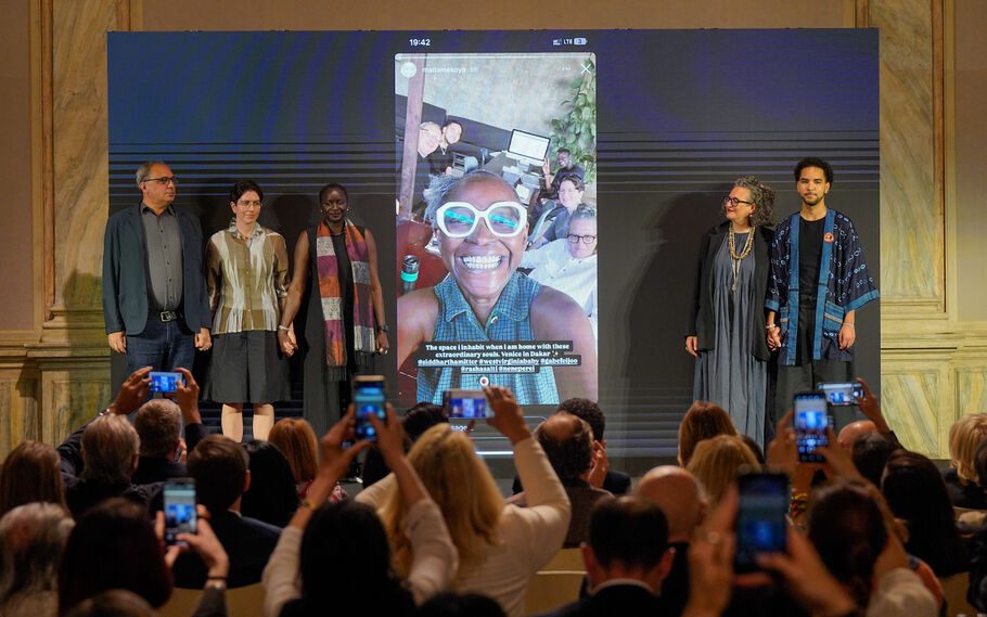 A group of five people (Venice Biennale 2026 curatorial team) standing on stage at a press conference with a projection of Koyo taking a selfie with a big smile showing between them.