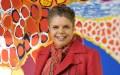 A 60-year-old First Nations woman with short gray hair smiling at the camera. She is wearing a red leather jacket and patterned neck scarf, standing in front of a vibrant artwork with large circles in red, orange, blue and yellow colours.