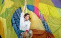 A circus performer appears to be rehearsing. They are hanging with one arm holding a thin rope and legs bent 90 degrees, against the backdrop of a large textile with blocks of neon yellow, blue, pink and purple colours.