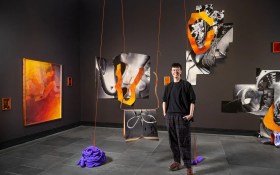 Jack Ball (A tall figure with short cropped hair smiling, wearing a black tshirt and pants) standing next to their large-scale installation featuring blurry photographs hanging from the ceiling and a purple pile of fabric to their right.