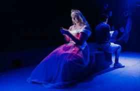 In blue light, a young man in a dress is looking at his phone. He's back to back with another man.