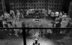 A black and white still from a video work depicting a single lone figure lying on their side in the middle of a concrete pen, with a crowd of blurry people walking in a large circle around them. A single onlooker is standing to the bottom of the image.