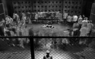 A black and white still from a video work depicting a single lone figure lying on their side in the middle of a concrete pen, with a crowd of blurry people walking in a large circle around them. A single onlooker is standing to the bottom of the image.