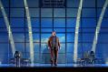 British countertenor James Laing as the Refugee in State Opera South Australia's 2025 production, 'Flight'. A distressed-looking man dressed in slightly dishevelled, mostly brown clothing, stands in an airport terminal between two rows of uncomfortable-looking metal and plastic seats. The blue-lit panelled wall behind him evokes the bars of a prison cell, while four curving beams of light projected against the walls increase the impression of this man being trapped or avoiding the spotlight.