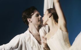 A man and female ballet dancer in white holding onto each other. She has one leg lifted high.