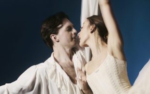A man and female ballet dancer in white holding onto each other. She has one leg lifted high.