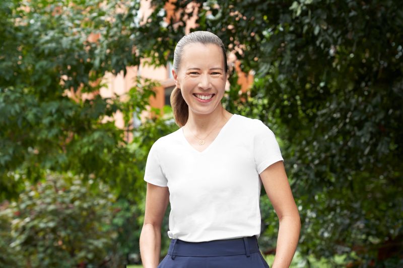 A young Caucasian woman with slicked back hair in a pony tail, standing in front of trees in a white V neck T shirt and blue skirt with her hands in the pockets. She is smiling at the camera.