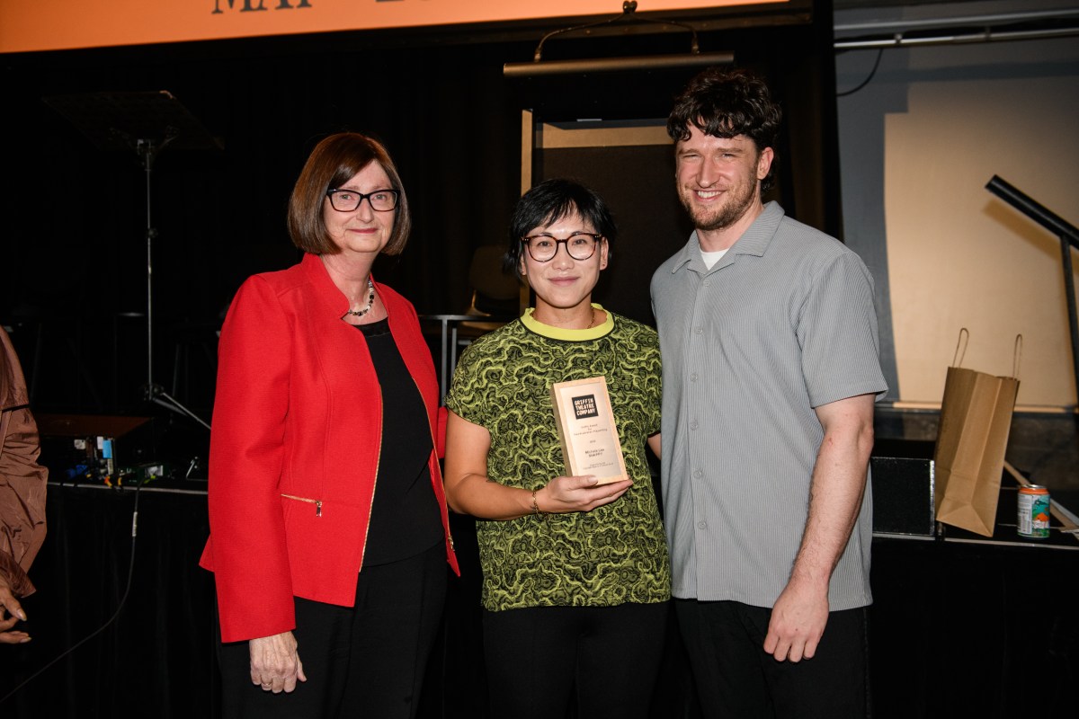 A photo of Michele Lee, a middle-aged Asian woman holding a trophy, with Kate Harrison, a middle-aged Caucasian woman, to her right and Declan Greene, a younger Caucasian man, to her left. They appear to be standing below the stage, facing and smiling for the camera.