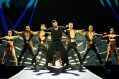 A Latin male pop singer on stage in black with a line-up of black clad dancers behind him. Ricky Martin.