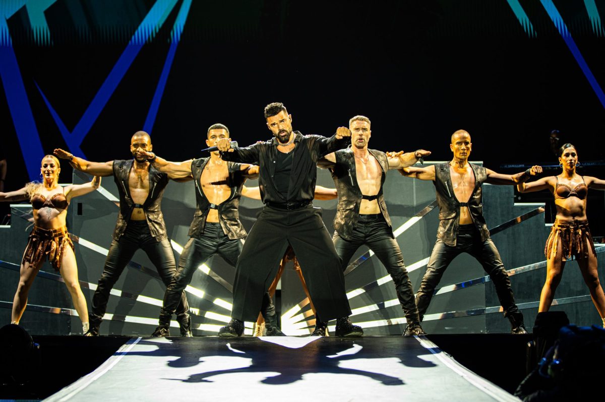 A Latin male pop singer on stage in black with a line-up of black clad dancers behind him. Ricky Martin.