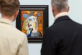 A colourful cubist artwork of the same woman seen from multiple perspectives, painted in shades of white, yellow, orange and blue. The painting is framed by the blurred bodies of two men who are looking at the artwork: the photo is taken over their shoulders. The painting is part of the exhibition 'Cézanne to Giacometti:' at the National Gallery of Australia.