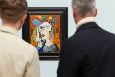 A colourful cubist artwork of the same woman seen from multiple perspectives, painted in shades of white, yellow, orange and blue. The painting is framed by the blurred bodies of two men who are looking at the artwork: the photo is taken over their shoulders. The painting is part of the exhibition 'Cézanne to Giacometti:' at the National Gallery of Australia.