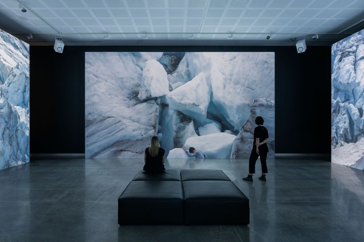 Two people are watching a large video installation in a gallery. The work shows shapes of glaciers, part of an environmental artwork called 'End & Being' by Jacobus Capone.