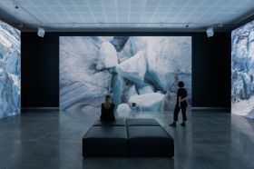 Two people are watching a large video installation in a gallery. The work shows shapes of glaciers, part of an environmental artwork called 'End & Being' by Jacobus Capone.
