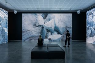 Two people are watching a large video installation in a gallery. The work shows shapes of glaciers, part of an environmental artwork called 'End & Being' by Jacobus Capone.