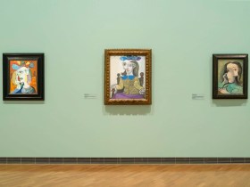 A picture of a wall at the National Gallery of Australia, featuring three modernist artworks. The wall is green. The floor is pine, and the three artworks are small in the frame of the photo. They show abstract works in bold colours.
