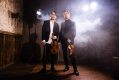 Classical musicians like Australian duo TwoSet Violin are finding thousands of fans on TikTok. The photo shows two younger Asian-Australian men, both formally dressed and holding violins by their sides in their left hands, and violin bows in their left. The two men are standing in a wooden studio surrounded by light and a single, standing spotlight illuminates the scene.