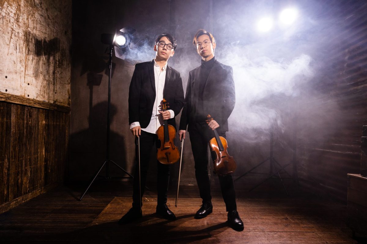 Classical musicians like Australian duo TwoSet Violin are finding thousands of fans on TikTok. The photo shows two younger Asian-Australian men, both formally dressed and holding violins by their sides in their left hands, and violin bows in their left. The two men are standing in a wooden studio surrounded by light and a single, standing spotlight illuminates the scene.