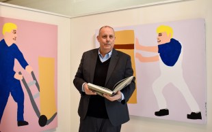 A bald man carrying a book standing in front of some artworks.