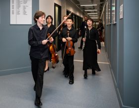 Four smiling young people from a range of cultural background, and all dressed in black and holding orchestral instruments, walk down a grey institutional hallway. They are members of the Australian Youth Orchestra's 2024 Young Symphonists program.