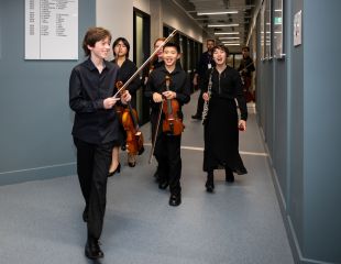 Four smiling young people from a range of cultural background, and all dressed in black and holding orchestral instruments, walk down a grey institutional hallway. They are members of the Australian Youth Orchestra's 2024 Young Symphonists program.