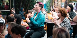 In the centre of the crowd of people is a woman with short pink hair and earrings wearing an aqua leather jacket.