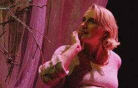A fair-skinned woman with wavy blonde hair bites one nail as she contemplates a budding tree branch. The scene is lit with a soft, warm light. A scene from the play 'Ripening' by Citizen Theatre.