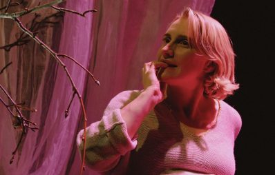 A fair-skinned woman with wavy blonde hair bites one nail as she contemplates a budding tree branch. The scene is lit with a soft, warm light. A scene from the play 'Ripening' by Citizen Theatre.