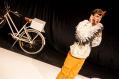 A pushbike on the left of the stage. On the right is a person dressed as a stork in yellow pants and a white ruffly top. A scene from Antipodes Theatre Company's 'Storked' at Theatre Works.