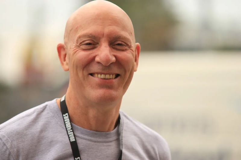 Clive Miller, Support Act CEO. A fair-skinned male-presenting person with a completely shaved head smiles at the camera. He wears a grey T-shirt and a black lanyard with the words 'Support Act' printed on it in white is included around his neck.