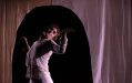 A dancer with their back bent and arms slightly outstretched, looking towards the right hand side. They are framed behind a curtain on stage with a dome-like opening.