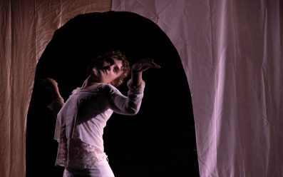 A dancer with their back bent and arms slightly outstretched, looking towards the right hand side. They are framed behind a curtain on stage with a dome-like opening.